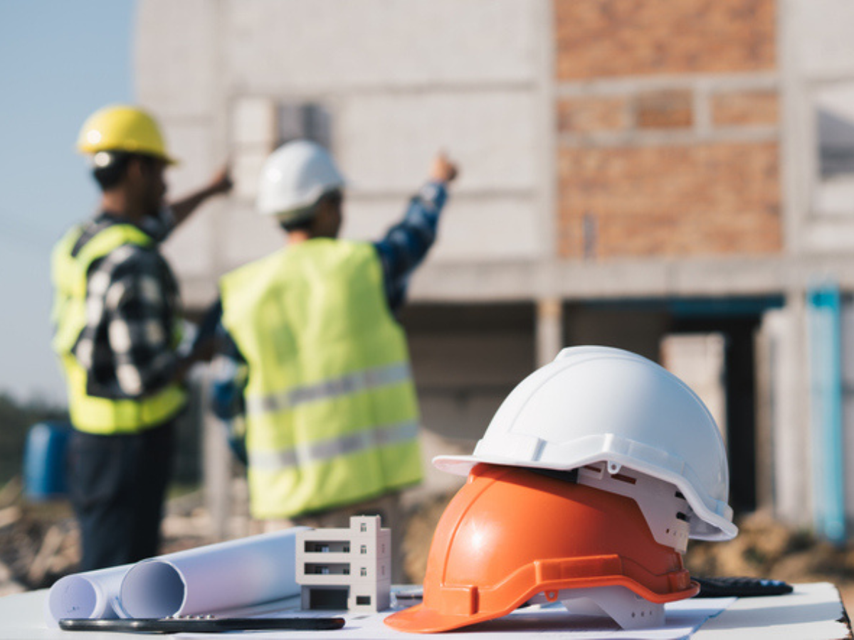 Hard hats and blueprints in the foreground with construction workers discussing project risks and insurance plans.