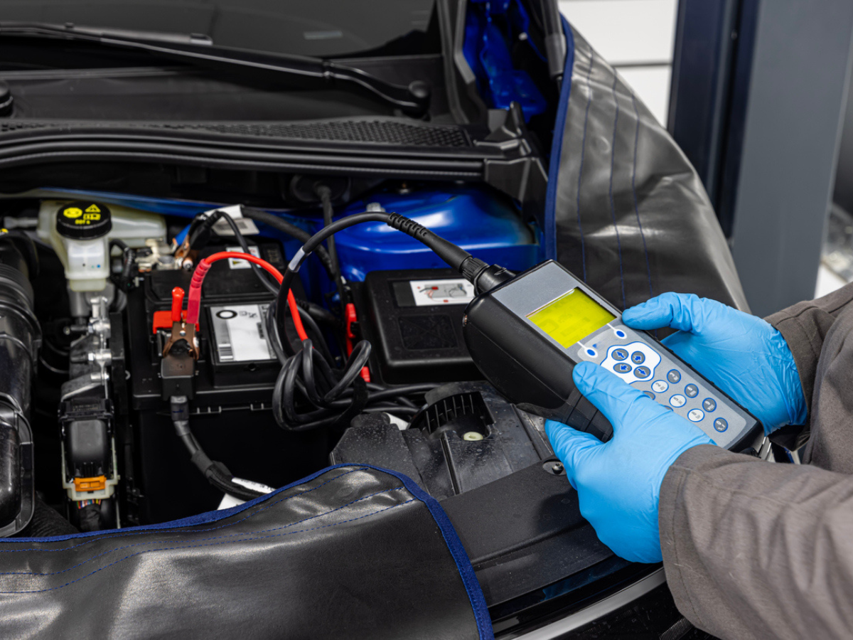 Gloved technician testing a car battery with a handheld digital analyzer under the open hood