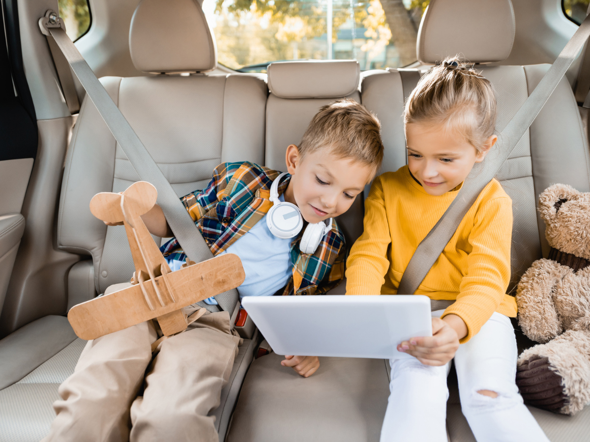 Two children wearing seat belts while using a tablet in the back seat of a car.
