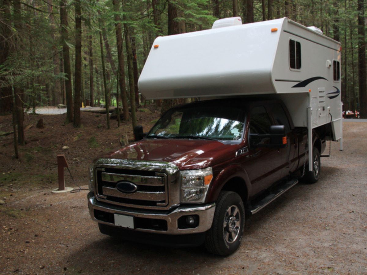 Slide-in truck camper mounted on a maroon pickup parked at a forest campsite