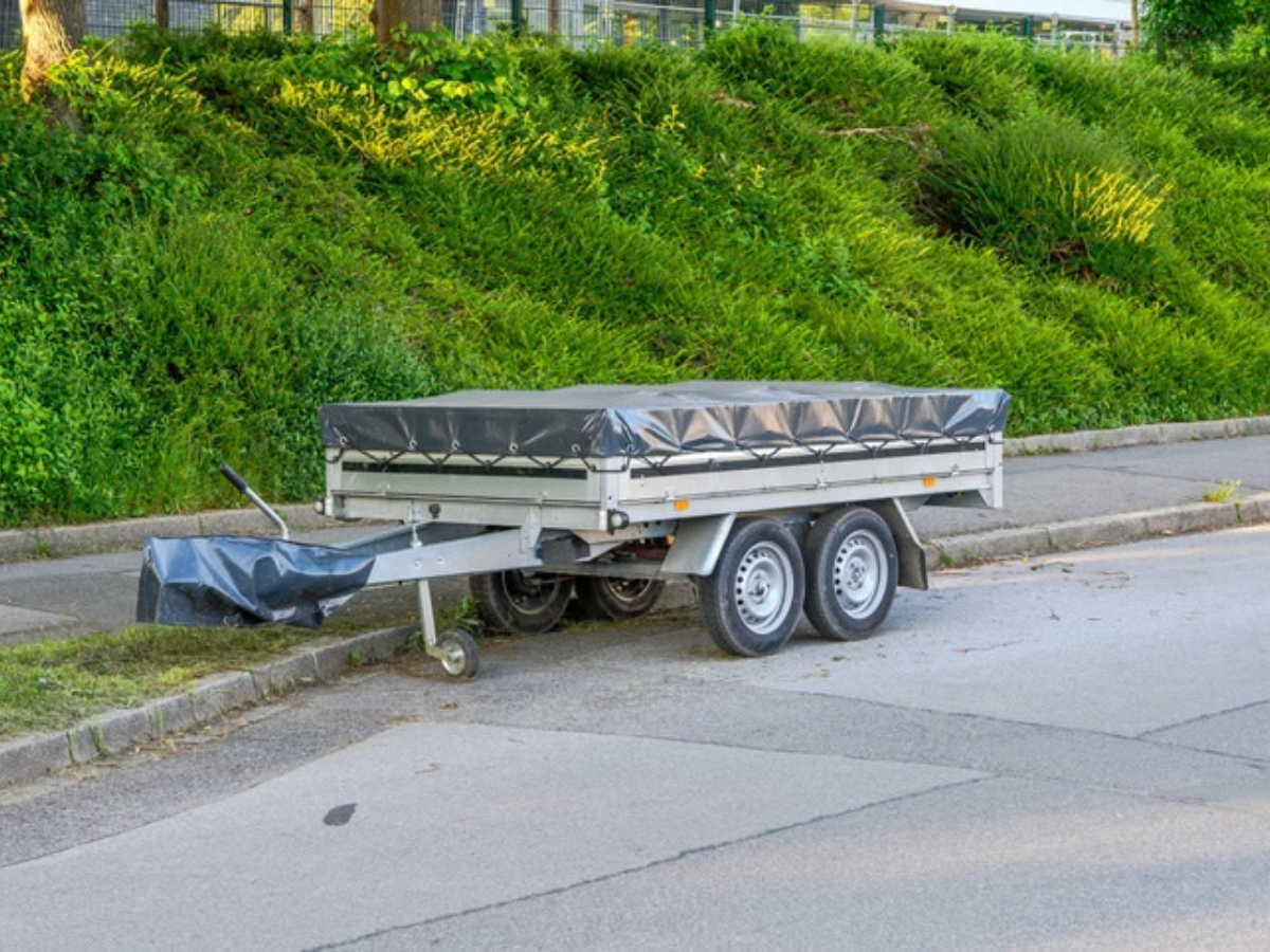 A covered utility trailer parked curbside next to lush greenery, illustrating how to safely store and insure your towed equipment.