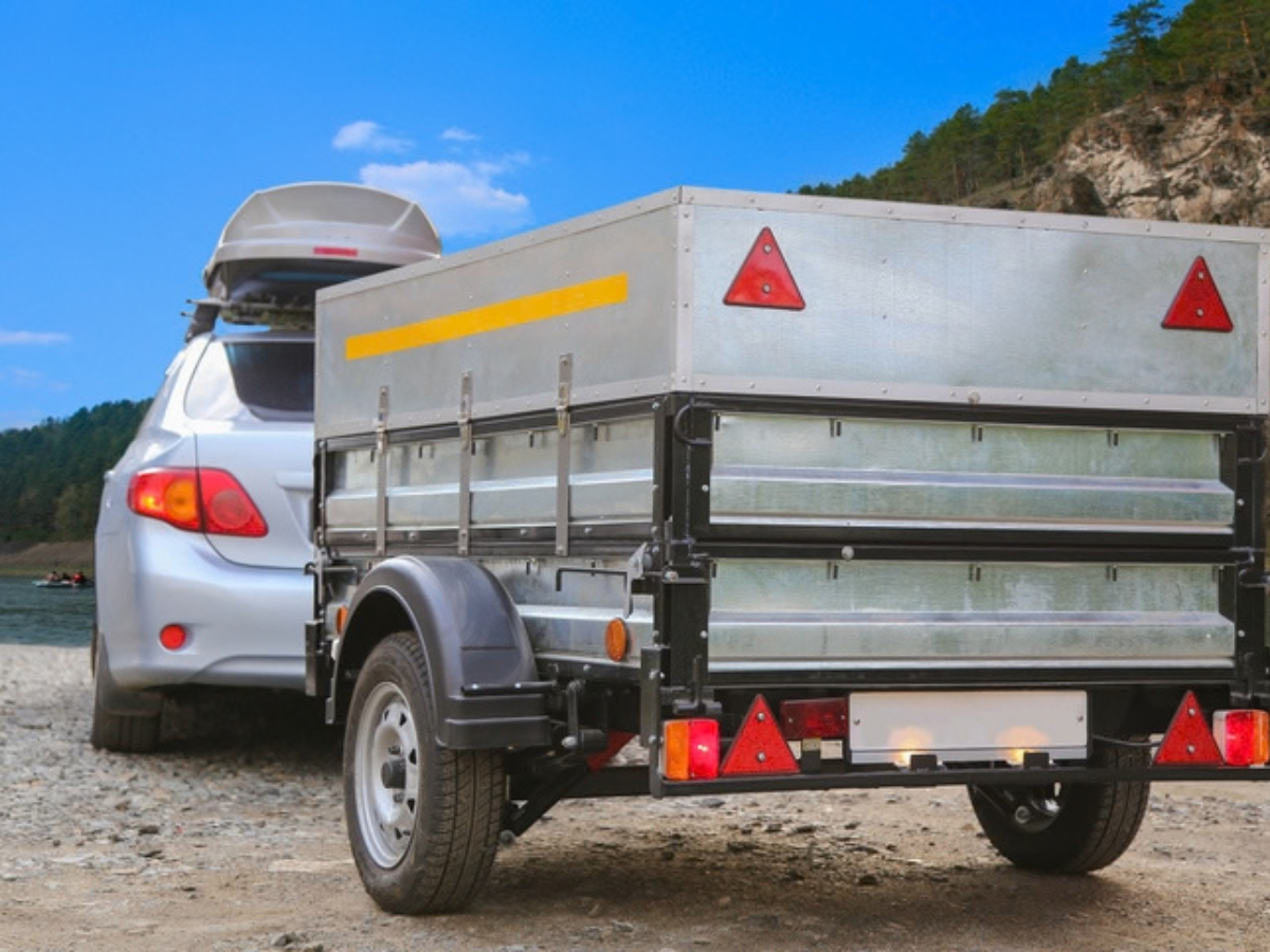 A silver car towing a cargo trailer parked near a scenic body of water, demonstrating coverage needed for road trips and hauling.