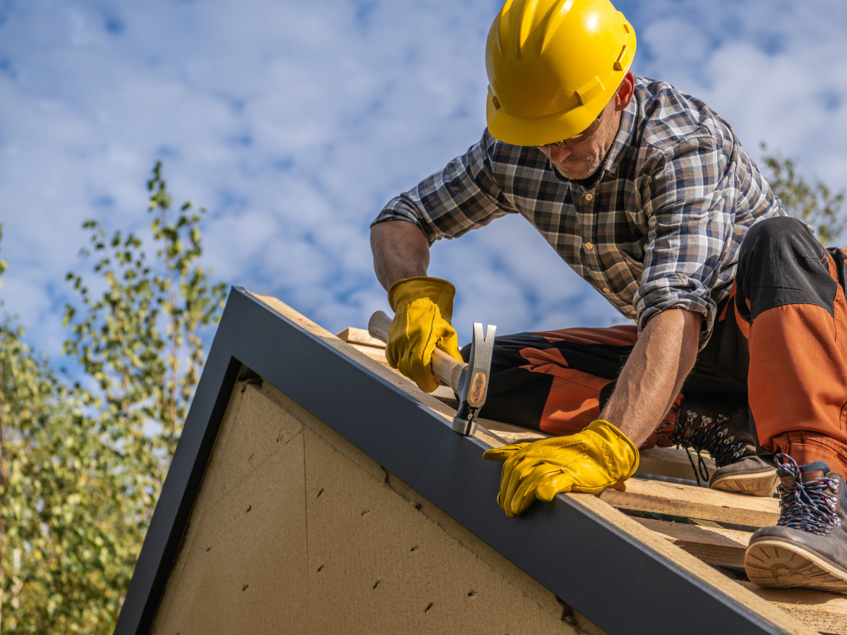 Construction worker wearing safety gear installs roof trim with a hammer on a residential building under clear skies.