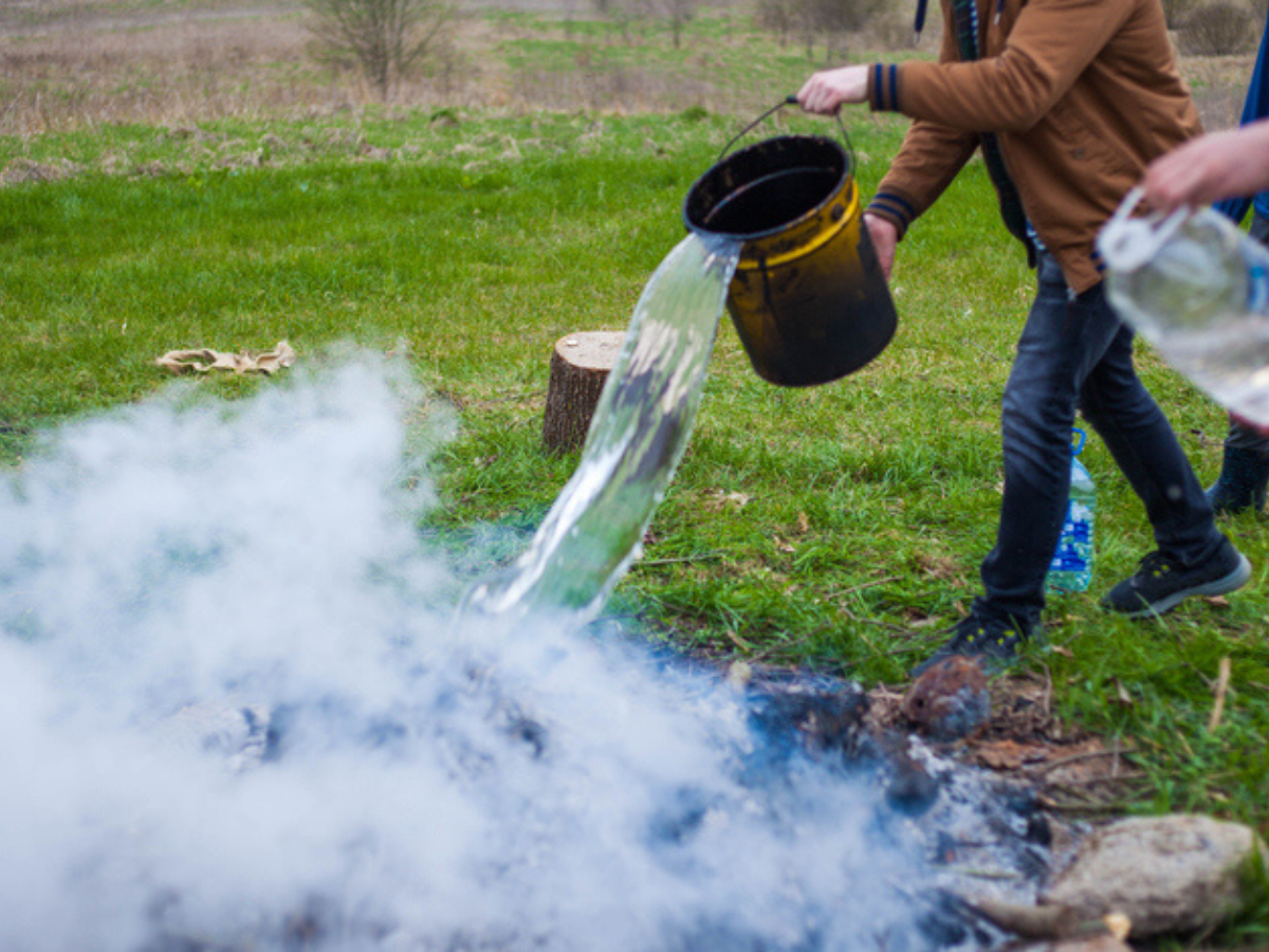 Two individuals pour water over a smoldering fire pit, demonstrating proper fire safety practices by fully extinguishing the flames to prevent accidental wildfires.