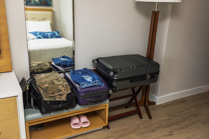 Packed suitcases with folded clothes in hotel room interior with mirror, wooden floor and pink slippers on shelf.