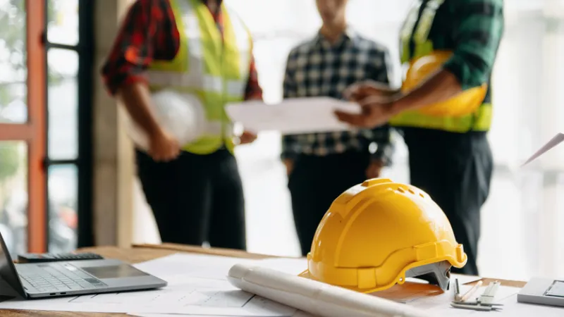 Contractors reviewing building plans while a hard hat and laptop sit on a work table