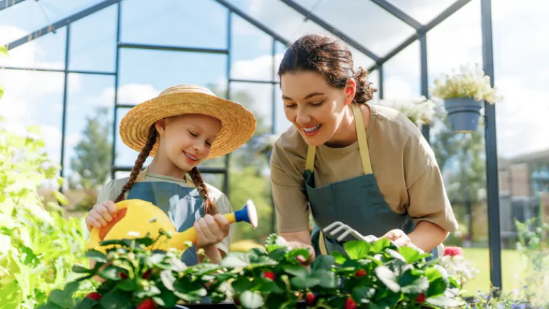 A woman and a young child smile while gardening together inside a greenhouse, watering and tending to green plants, representing family life and small-scale hobby farming.