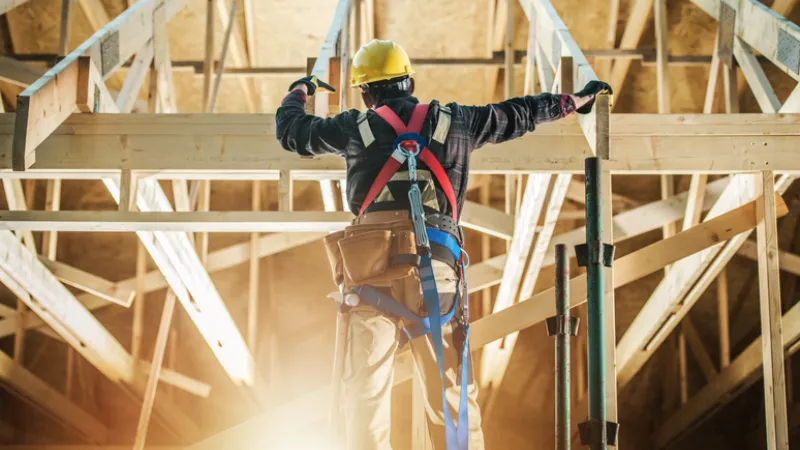 A construction worker wearing safety gear installing roof trusses on a building site protected by builders risk insurance
