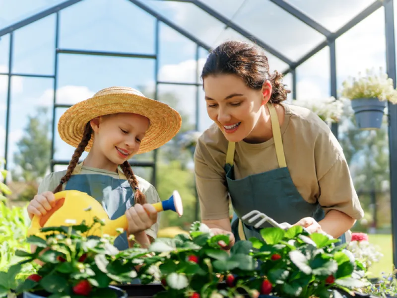 A woman and a young child smile while gardening together inside a greenhouse, watering and tending to green plants, representing family life and small-scale hobby farming.