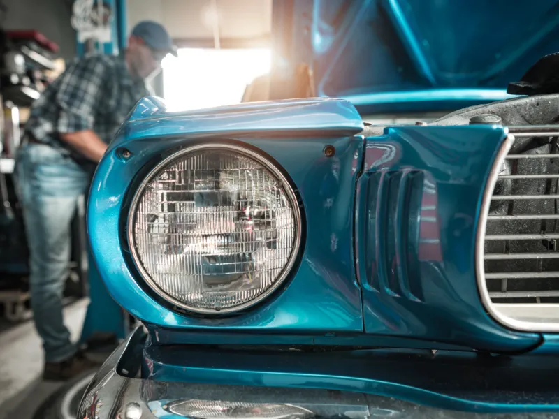 Close up of a restored vintage car headlight in a garage with a mechanic working in the background, representing classic vehicle restoration and insurance